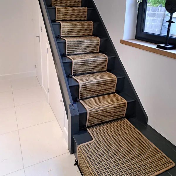 Black stair rods securing a textured woven seagrass runner on dark grey stairs.