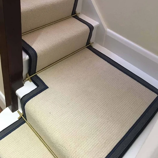 Brass stair rods securing a beige runner with a dark blue border on white stairs.