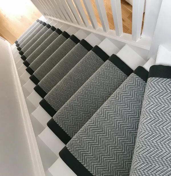Light grey herringbone stair runner with dark binding on white stairs.