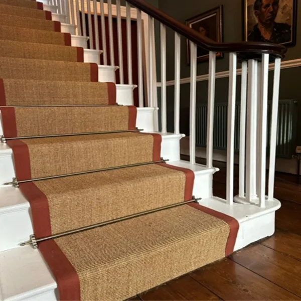 Metal stair rods securing a natural fiber runner with red border on white stairs.