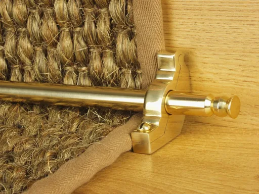 Polished brass stair rod securing a natural fiber runner with brown fabric binding.